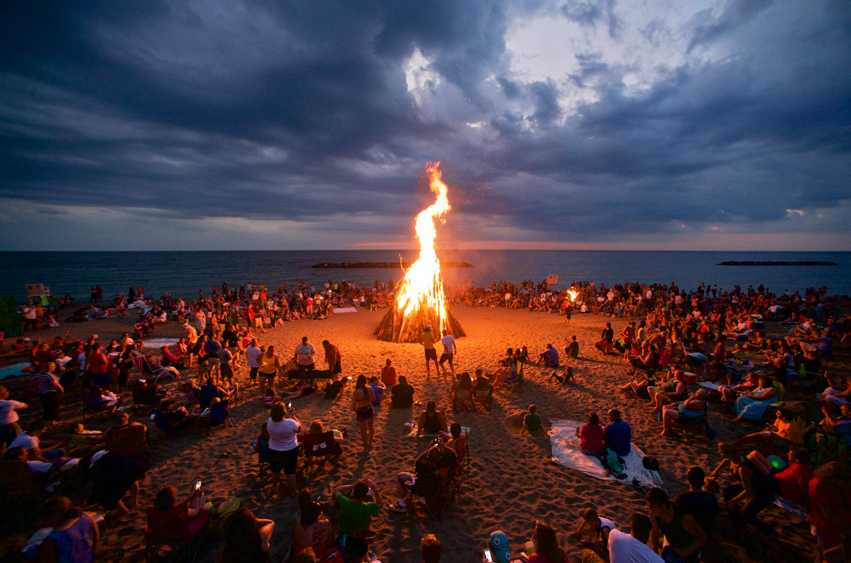 A large crowd gathers around a bonfire on the beach.